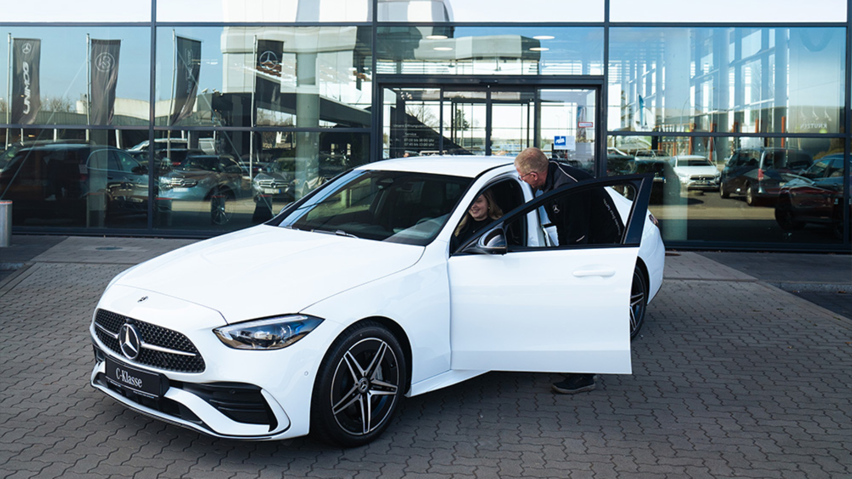 Weiße C-Klasse vor einem Mercedes-Benz Gebäude. Ein Mitarbeiter beugt sich zur im Fahrzeug sitzenden Kundin runter.