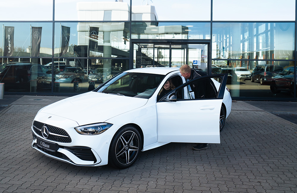 Weiße C-Klasse vor einem Mercedes-Benz Gebäude. Ein Mitarbeiter beugt sich zur im Fahrzeug sitzenden Kundin runter.